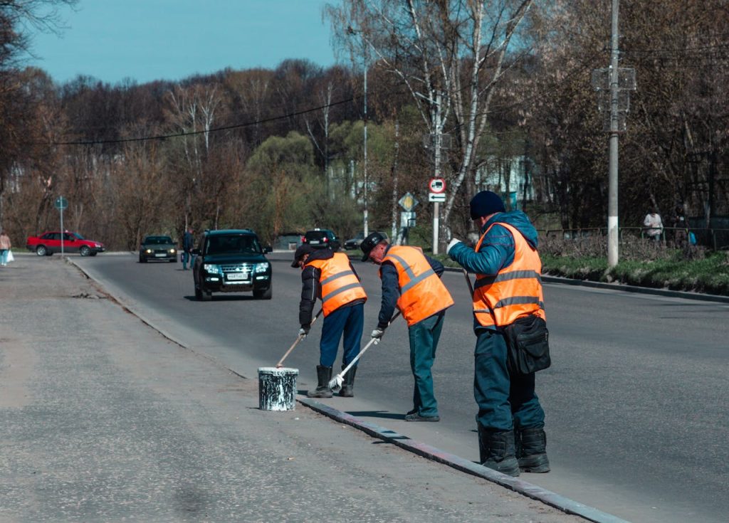 Three road workers in reflective vests paint street markings on a suburban road.